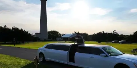 A white stretch limousine parked on a paved path with a red carpet leading to the open door, set against a backdrop of a lighthouse and green grass.