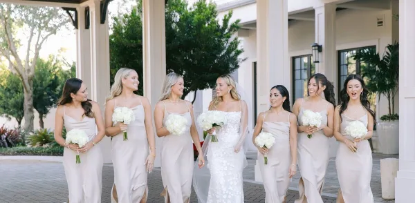 A bride and her bridesmaids in matching champagne dresses holding white bouquets, standing together at a wedding venue.