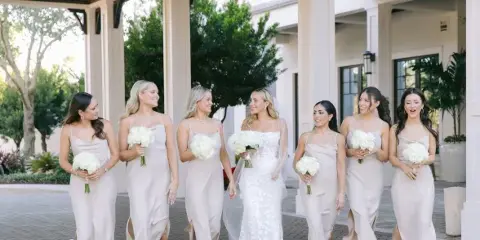 A bride and her bridesmaids in matching champagne dresses holding white bouquets, standing together at a wedding venue.