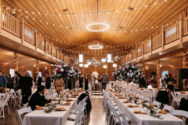 Interior view of a rustic wooden wedding venue with string lights, chandeliers, and long banquet tables set for a reception.