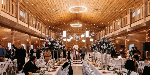 Interior view of a rustic wooden wedding venue with string lights, chandeliers, and long banquet tables set for a reception.