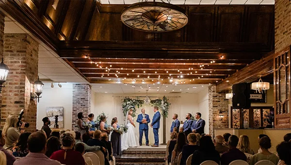 A wedding ceremony in progress inside a rustic venue featuring exposed brick walls, wooden ceiling beams, and a stained-glass skylight.