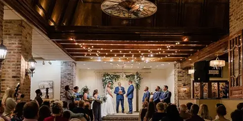 A wedding ceremony in progress inside a rustic venue featuring exposed brick walls, wooden ceiling beams, and a stained-glass skylight.