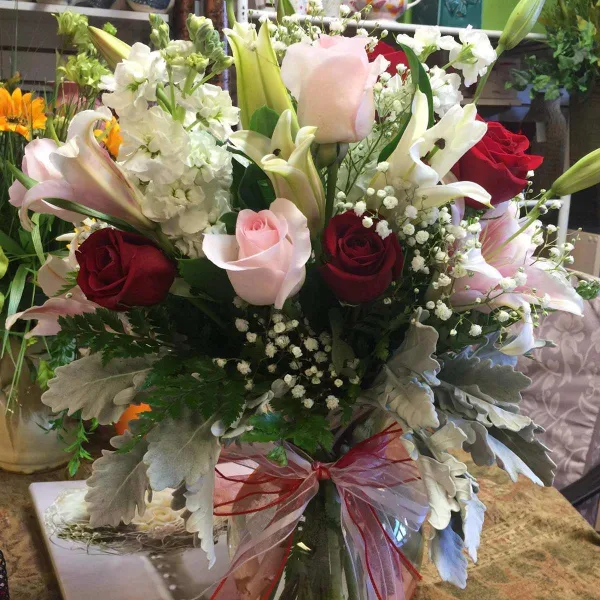 A floral arrangement with red and pink roses, white lilies, and baby's breath tied with a ribbon, displayed in a florist shop.