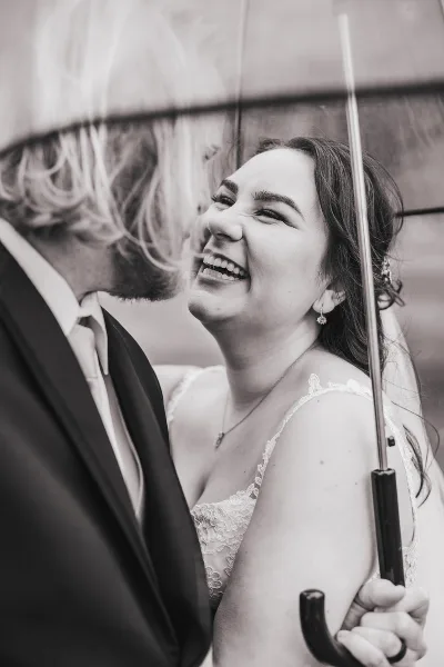A black and white photograph of a bride and groom laughing together while standing under an umbrella.