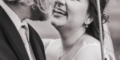 A black and white photograph of a bride and groom laughing together while standing under an umbrella.