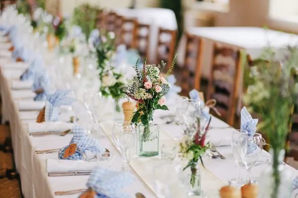 Styled event table with white linens, blue gingham napkins, glassware, and floral centerpieces.