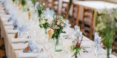 Styled event table with white linens, blue gingham napkins, glassware, and floral centerpieces.