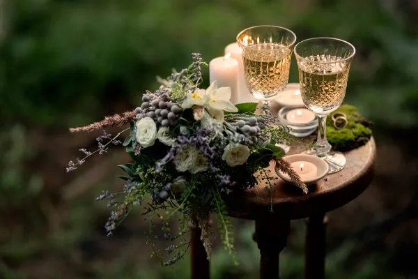 A rustic wedding table setting featuring a floral bouquet, lit candles, and two glasses of champagne on a wooden stool.