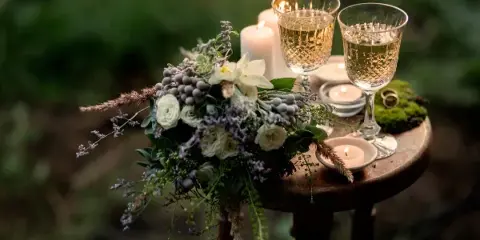 A rustic wedding table setting featuring a floral bouquet, lit candles, and two glasses of champagne on a wooden stool.