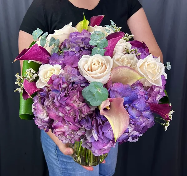 A person holding a large, handcrafted bouquet of white roses, purple hydrangeas, and calla lilies.