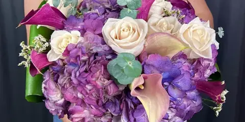 A person holding a large, handcrafted bouquet of white roses, purple hydrangeas, and calla lilies.