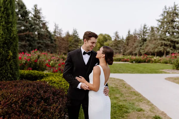 A bride and groom standing together in a garden setting, looking at each other and smiling.