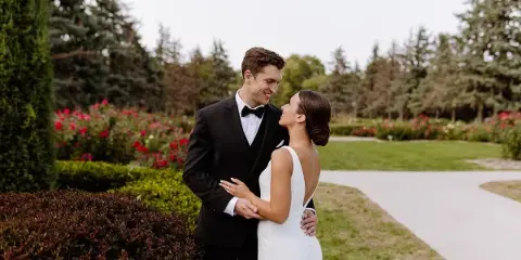 A bride and groom standing together in a garden setting, looking at each other and smiling.