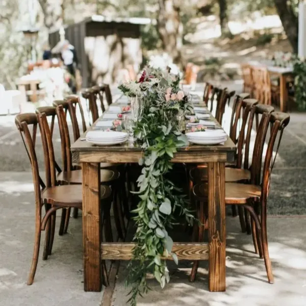 A long wooden farm table set with white plates and a floral runner, surrounded by wooden chairs.