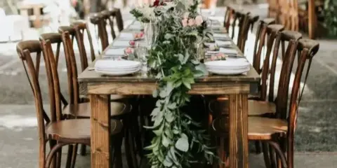 A long wooden farm table set with white plates and a floral runner, surrounded by wooden chairs.