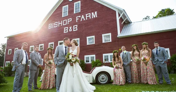 A wedding party posing in front of a large red barn with 'Bishop Farm B&B' painted on the side.