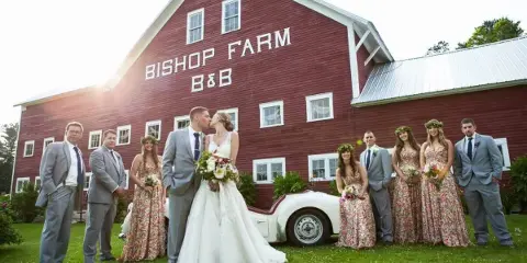A wedding party posing in front of a large red barn with 'Bishop Farm B&B' painted on the side.