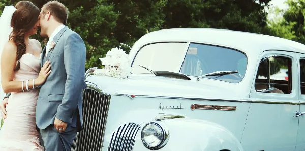 A bride and groom standing next to a vintage white Packard wedding car with a floral arrangement on the hood.