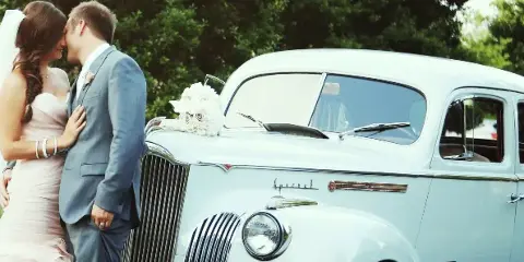 A bride and groom standing next to a vintage white Packard wedding car with a floral arrangement on the hood.