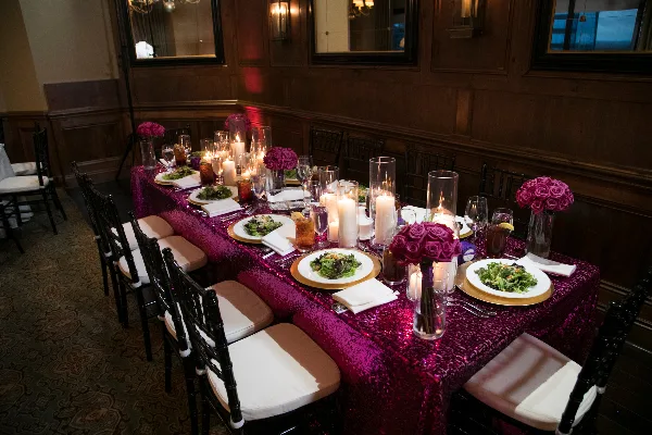 A long event table set with black Chiavari chairs, a magenta sequined tablecloth, and floral centerpieces in a wood-paneled room.