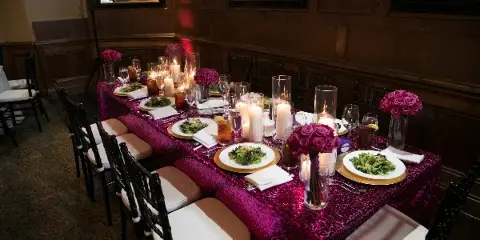 A long event table set with black Chiavari chairs, a magenta sequined tablecloth, and floral centerpieces in a wood-paneled room.