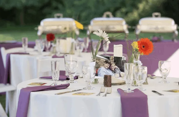 An outdoor catering table set with white linens, purple napkins, glassware, and silver chafing dishes in the background.