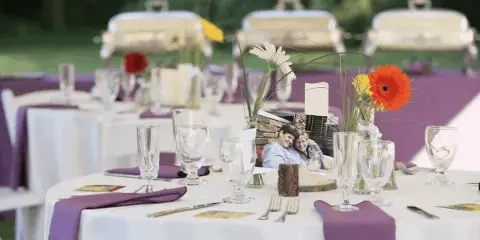 An outdoor catering table set with white linens, purple napkins, glassware, and silver chafing dishes in the background.