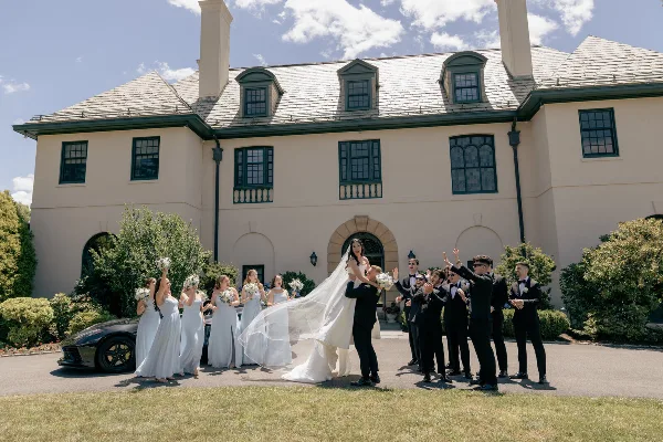 A wedding party, including the bride and groom and their attendants, posing outdoors in front of a large estate building.