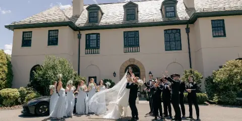 A wedding party, including the bride and groom and their attendants, posing outdoors in front of a large estate building.
