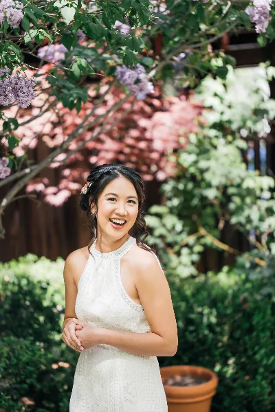 A smiling bride with styled hair and makeup standing outdoors in a white lace dress.