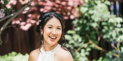 A smiling bride with styled hair and makeup standing outdoors in a white lace dress.