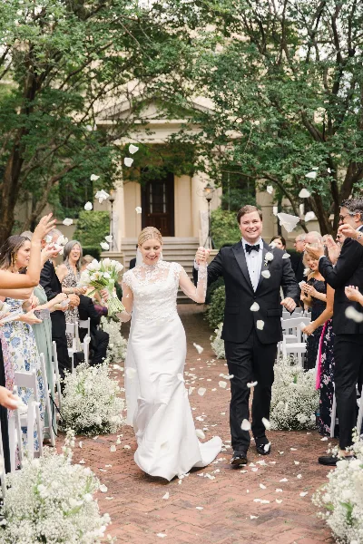 A bride and groom walking down a brick aisle lined with white flowers and guests during a wedding ceremony.