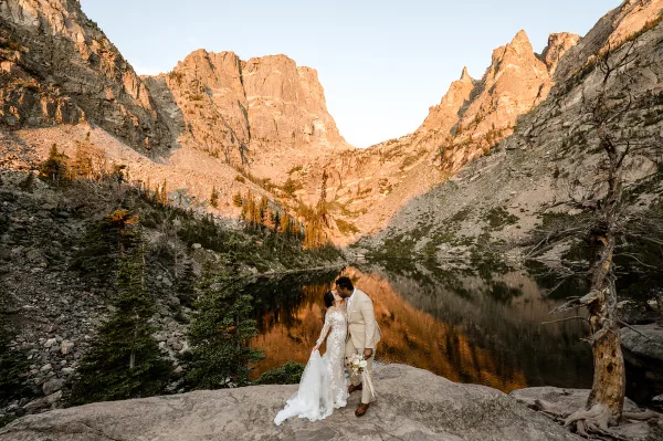 A bride and groom standing on a rocky ledge overlooking a mountain lake with steep peaks in the background.