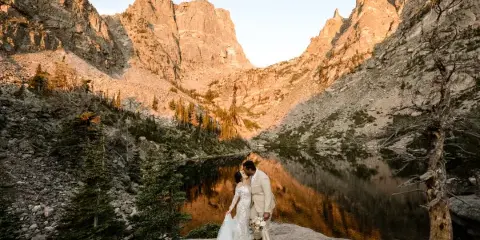 A bride and groom standing on a rocky ledge overlooking a mountain lake with steep peaks in the background.