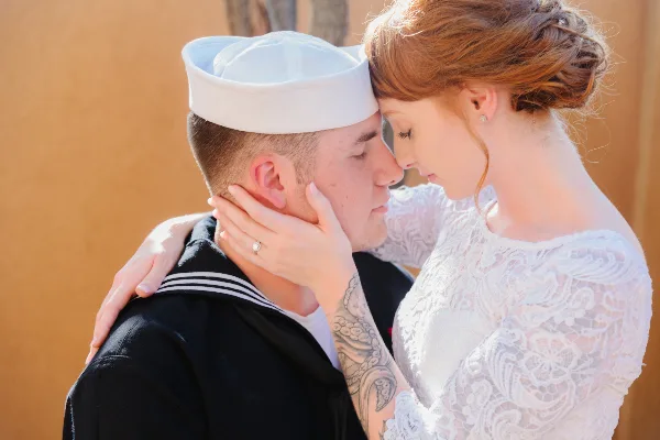 A bride in a white lace dress and a groom in a navy sailor uniform share an intimate moment, foreheads touching, with the bride's hand gently cupping the groom's face.
