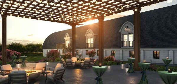 Outdoor wedding venue patio with a wooden pergola, string lights, lounge seating, and a large barn-style building with arched windows and balconies at sunset.