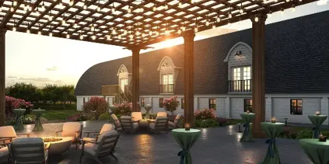 Outdoor wedding venue patio with a wooden pergola, string lights, lounge seating, and a large barn-style building with arched windows and balconies at sunset.