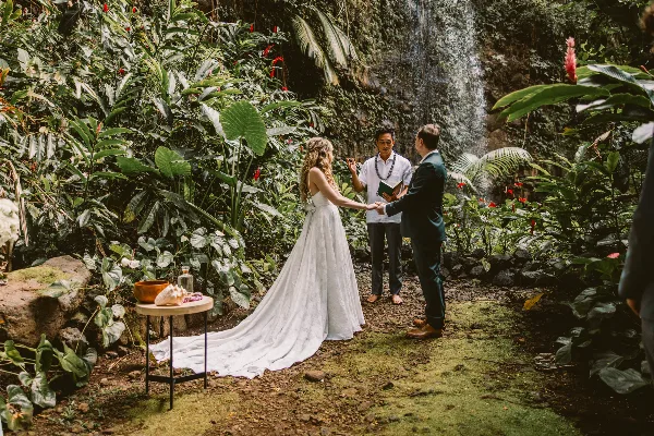 A wedding officiant stands between a bride and groom during an outdoor ceremony in a lush, tropical garden setting.