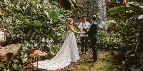 A wedding officiant stands between a bride and groom during an outdoor ceremony in a lush, tropical garden setting.