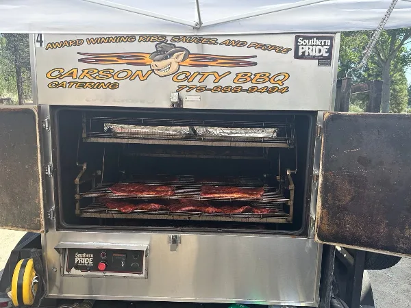 A commercial smoker branded with Carson City BBQ Catering, showing racks of ribs cooking inside.