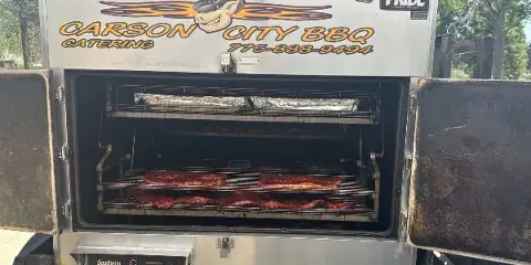 A commercial smoker branded with Carson City BBQ Catering, showing racks of ribs cooking inside.