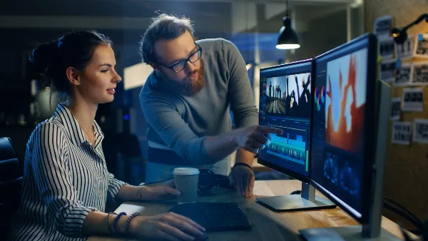 Two professionals reviewing video footage and editing timelines on dual monitors in a studio setting.