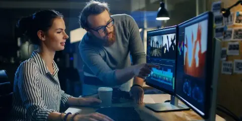 Two professionals reviewing video footage and editing timelines on dual monitors in a studio setting.