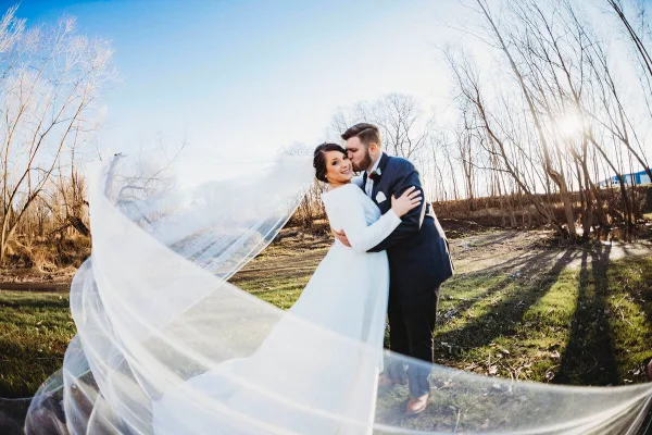 A bride and groom embracing outdoors with the bride's veil flowing in the wind.