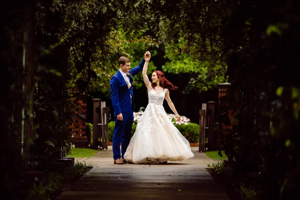 A bride in a white lace gown and a groom in a blue suit dancing on a stone pathway surrounded by greenery.