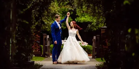 A bride in a white lace gown and a groom in a blue suit dancing on a stone pathway surrounded by greenery.