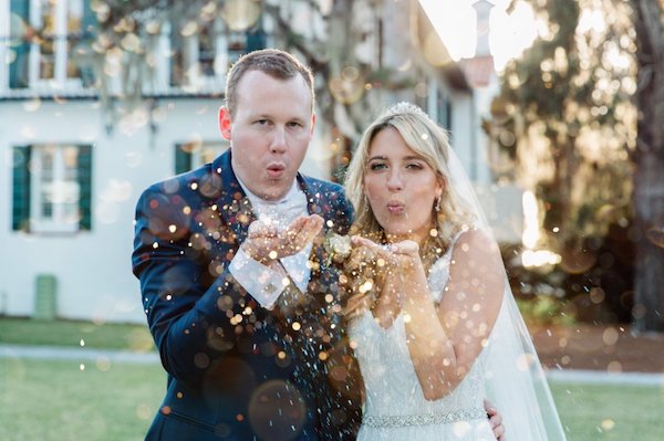 A bride and groom blowing gold glitter in front of a white building with green shutters.