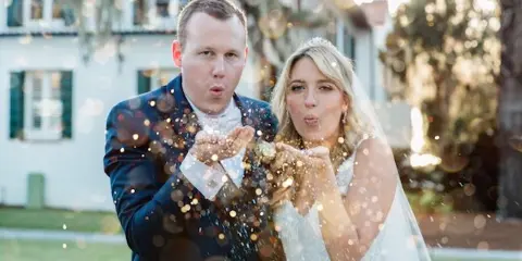 A bride and groom blowing gold glitter in front of a white building with green shutters.