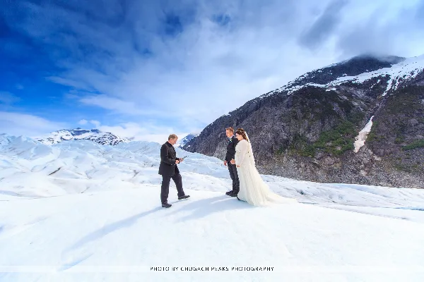 A bride and groom standing on a glacier during a wedding ceremony with mountains in the background.